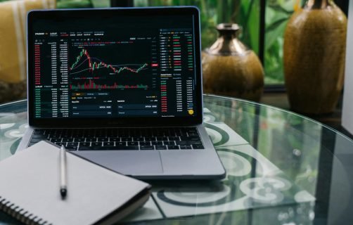 Laptop displaying cryptocurrency stocks and graphs on a glass table with a notepad.