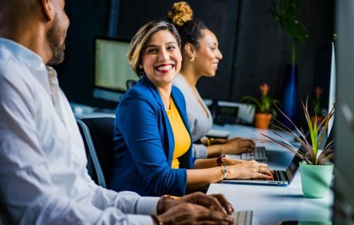 Three diverse professionals working and smiling at office desks, fostering teamwork and collaboration.