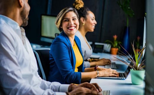 Three diverse professionals working and smiling at office desks, fostering teamwork and collaboration.