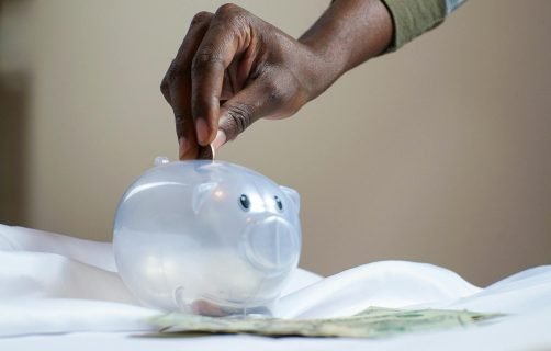 Close-up of a person's hand placing coins into a transparent piggy bank to save money.
