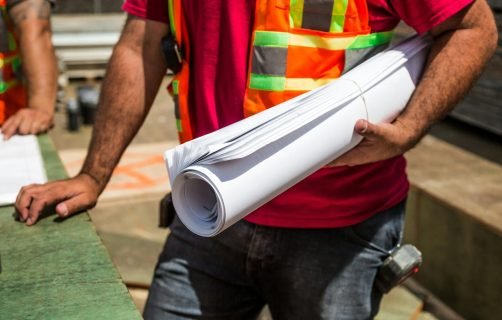 Construction workers in safety vests holding blueprints at a site during the day.