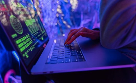 Close-up of hands typing on a laptop displaying cybersecurity graphics, illuminated by purple light.