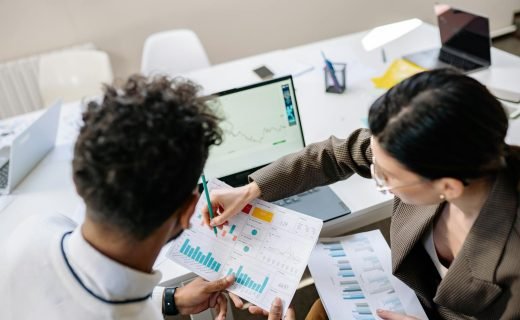 Two colleagues in a meeting room discussing financial charts and graphs on a laptop and paper.