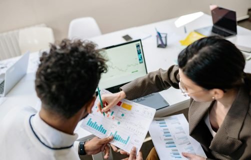 Two colleagues in a meeting room discussing financial charts and graphs on a laptop and paper.