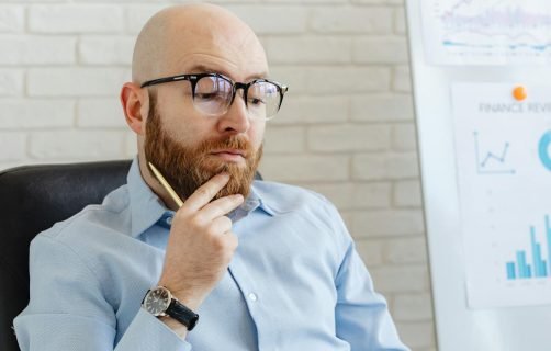 Bearded man in blue shirt contemplating financial data on whiteboard indoors.